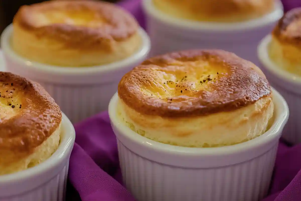 Five golden soufflés in white ramekins, garnished with freshly cracked black pepper, placed on a purple napkin for an elegant presentation.