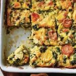 Baked Easter breakfast casserolein a white dish, sliced into squares with one corner piece removed, showcasing tomatoes, greens, and melted cheese.